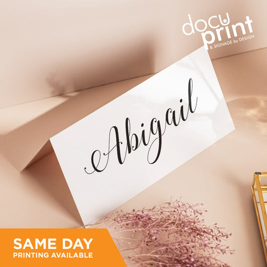 A folded place card with 'Abgaili' written in cursive script, presented on a beige background with a small floral arrangement to the side, custom designed and printed by Docuprint in Fremantle, Perth, Australia