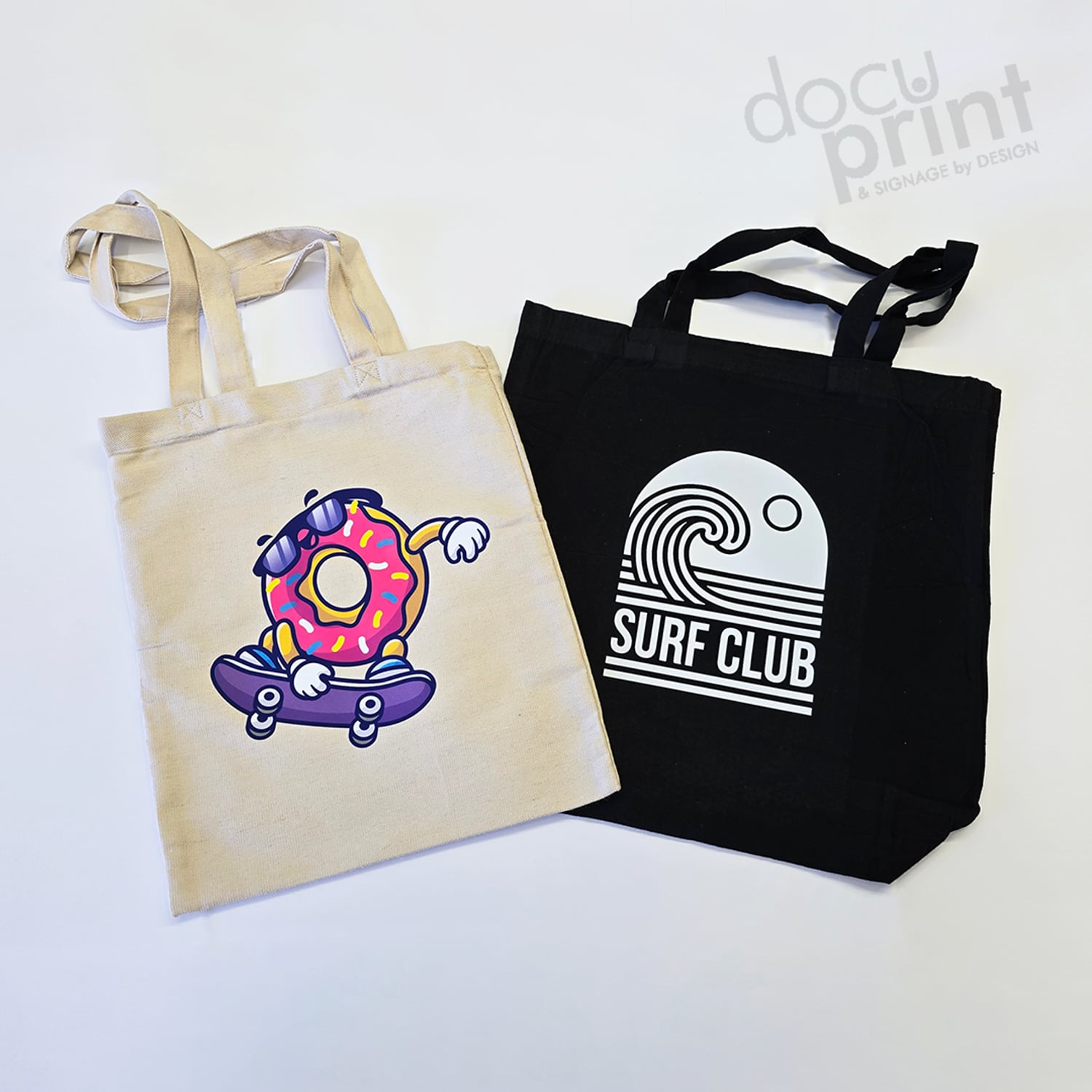 white cotton tote bag with a donut riding a skateboard on it, next to a black cotton tote bag that has a graphic wave and says 'surf club'. Custom designed and printed by Docuprint Fremantle, in WA.